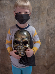 Obraz premium Funny blond kid with scary skeleton mask in his hands in black medical mask to protect against influenza viruses in crowded places. Chinese inscription on the wall background, Wuhan coronavirus