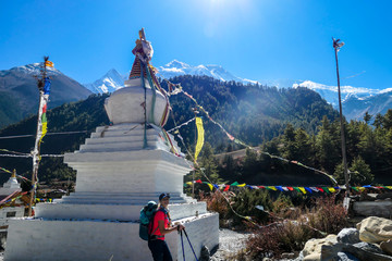 A woman trekking along a small white pagoda, Annapurna Circuit Trek in Nepal. She is enjoying the view and trek. Snow caped mountains in the back. Bright and sunny day.Meditation and peace of mind