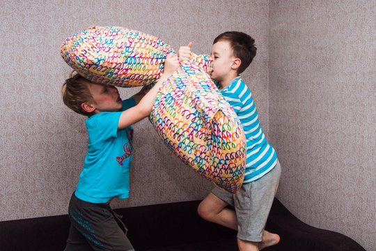 Two Brothers Of A European Boy Fight With Pillows On The Bed. A Merry Duel Of Children. Fight At Home Between Friends, War On Purpose.