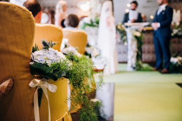 Wedding ceremony area, flowers composition with greenery and ribbons on the yellow chairs, bride and a groom on a background