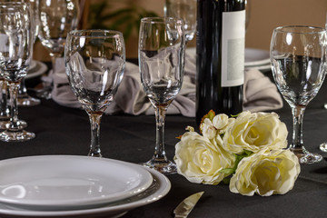 Platos blancos con copas en primer plano y botella de vino en mesa negra con rosas color crema
