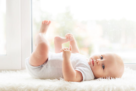 a small child about 6 months old lies by the window on a white blanket and catches its legs