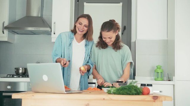 Two young caucasian women dancing while cooking and talking with friends via the internet. Friends are having fun while cooking together.