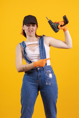 Builder girl in denim overalls and a black cap holds a drill and a screwdriver in her hands and...