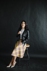 portrait of a young dark-haired girl on a dark background sitting on a bar stool with dark leather jacket and skirt. Photography of a young beautiful girl in the Studio