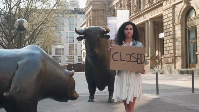 Frankfurt, Germany, April 8, 2020 Young women against corona virus holding sign with closed in front of bull and bear stock market exchange shot in 4k