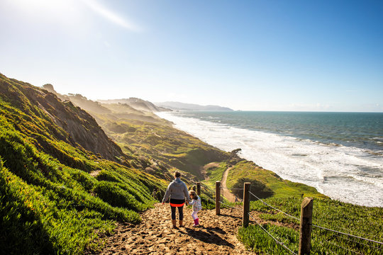 A young girl and her mother hiking together on a sunny day near the Pacific Ocean with warm clothes.