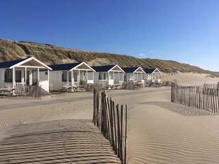 Rental beach houses neatly in row in beach against dunes with table in front and fences, with late afternoon shadows