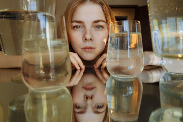 The girl looks at the camera. The face of a Girl with yellow eyes is reflected in the mirror. She has beautiful facial features. There are cans of coke around. There are bubbles in the water.