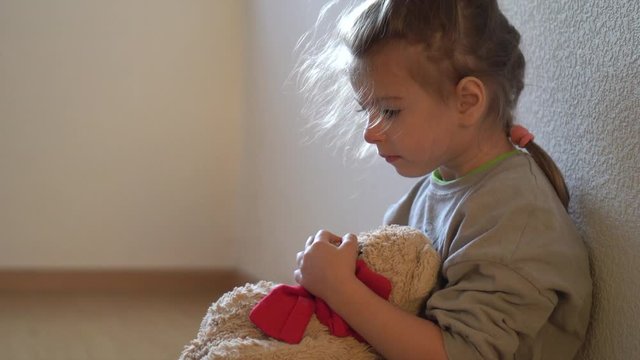 Abandoned child. Little girl beside the wall by the window alone with teddy bear. Sad difficult childhood.