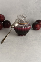 Glass jar of plum jam with spoon and berries on textured white stone background. Homemade marmalade for breakfast. Copy space. 