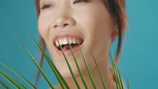 A beautiful young asian woman is looking through a plant leaf to the camera while touching it over blue wall background