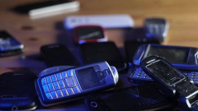 Old broken scratched and dusty cell phones lie in a pile on the table in blue neon light. Analog communication in the office. Modern wireless technology. Close-up of an old and outdated mobile phone.