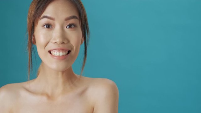 A happy positive young asian woman is pointing to the side with her finger over blue wall background