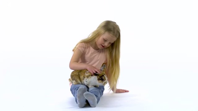 Little girl with long blonde hair is stroking fluffy threecolored rabbit at white background. Slow motion