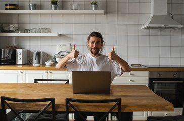 Excited businessman feeling happy about successful project. Man holding thumbs up in the air and looking at laptop while sitting on a kitchen. Happy freelancer, student working at laptop at home.