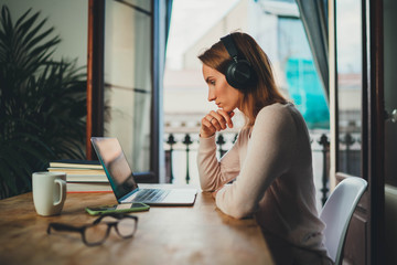 Concentrated student girl learning online having video call via laptop computer sitting at home interior near open balcony, female entrepreneur working remotely from home office using modern computer