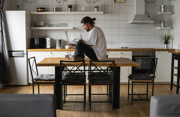 Freelancer working from home sitting on a top of the kitchen table and using laptop. Bearded man working with a laptop and reading news. Successful self entrepreneur working at his modern home.