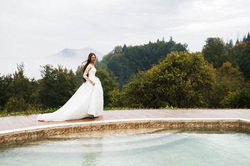 Young woman with long hair dressed in wedding dress walks by the pool outdoors in autumn.