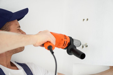 Young professional worker with drill making a hole in white wall.