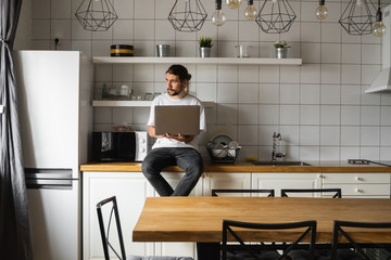 Freelancer working from home sitting on a kitchen worktop and using laptop. Bearded man working with a laptop and reading news. Handsome successful self entrepreneur working at his modern home.