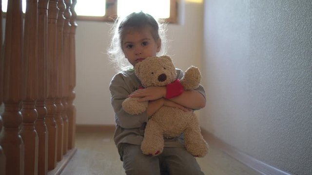 Abandoned child. Little girl beside the wall by the window alone with teddy bear. Sad difficult childhood.