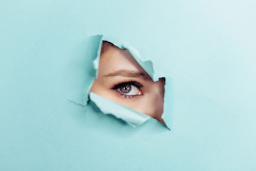 Eye of the beautiful woman looking through hole in blue paper. Spy eye watching through hole.