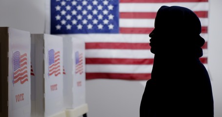 MS silhouetted African American man in a hoodie looking at voting booths, with US flag on wall...