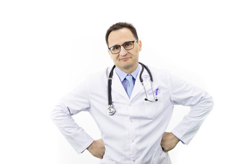 The therapist with glasses in a white coat with a stethoscope stands on a white background and looks at the camera. Hands on hips. Medical worker. Serious look. Portrait of a doctor. Smirk. Close-up.