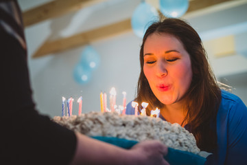 Young caucasian brunette woman blowing out candles on a birthday cake with candles. Tasty birthday present for a girl at a indoor party.