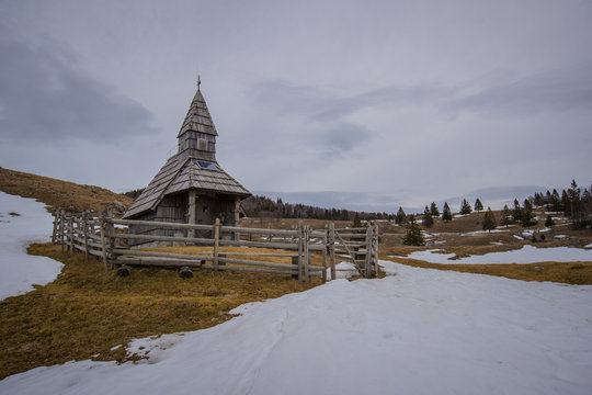 Small wooden church surrounded with fence on a mountain plateu of biba planina in Slovenian Alps. Early spring with patches of snow around still visible.