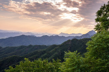 Beams of evening sunlight spill through the peaceful clouds over the mountains surrounding the Great Wall of China near Beijing, China.
