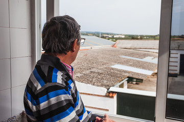 Senior man leaning out the terrace window at home looking at the street. Selective focus.