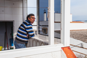 Senior man leaning out the terrace window at home looking at the street. Selective focus.