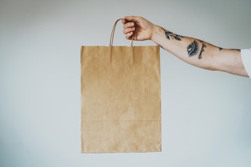 cropped shot on a hand with tattoos that holding craft paper package with empty space for your logo or design, mock-up of shopping bag with handles. White wall background - 337489827