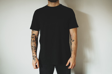 young attractive male hipster with a beard and tattoos, dressed in a black blank t-shirt, posing on a white wall background. Empty space for you logo or design. - 337489871