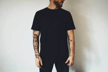 young attractive bearded man with tattoos, dressed in a black blank t-shirt, posing on a white wall background. Empty space for you logo or design. - 337489890