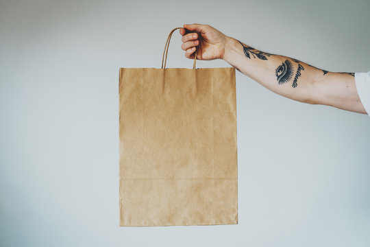 cropped shot on a hand with tattoos that holding craft paper package with empty space for your logo or design, mock-up of shopping bag with handles. White wall background