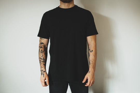 young attractive male hipster with a beard and tattoos, dressed in a black blank t-shirt, posing on a white wall background. Empty space for you logo or design.