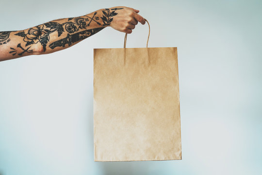 cropped photo on a hand with tattoos that holds blank craft paper package, bag mock-up. Empty space for design. White wall background