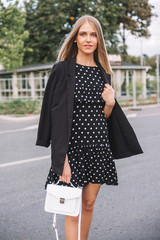 Very Beautiful young woman with long hair in a black short dress, a jacket with a small white hand bag. Posing Summer evening on a city street.