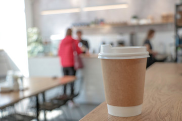 Mock up with a cup of coffee. A glass is on the table in a cafe, selling coffee in the background. Coffee to go. Light walls. Wooden table. Cafe. Disposable cup