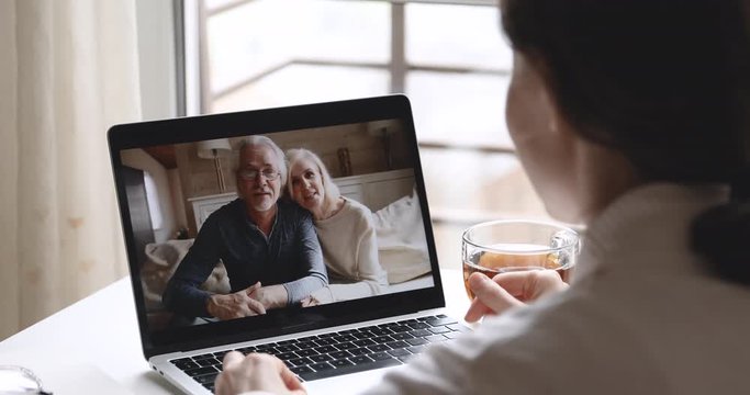 Over shoulder closeup view of young woman daughter video calling old senior parents talking with grandparents mom dad in webcam conference chat app on laptop screen on table. Family videocall concept.