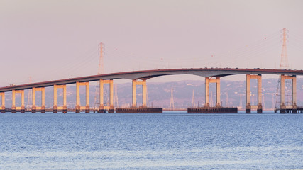Sunset view of San Mateo Bridge connecting the Peninsula and East Bay in San Francisco Bay Area, California; Electricity towers and power lines visible behind it