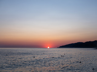 People are swimming in the Black sea  during sunset. A red disk of the sun goes under the water. On the right there is a silhouette of the root of the mountain.