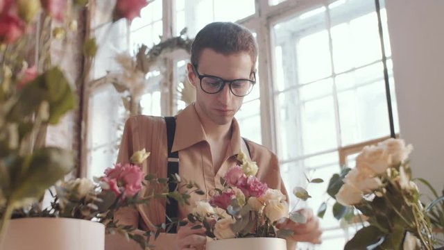 Young handsome male florist in glasses and apron arranging beautiful flower composition in hatbox, then leaning on table, looking at camera and smiling during workday in flower shop