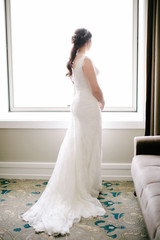 The bride poses facing back. He looks out the window. Indoor.