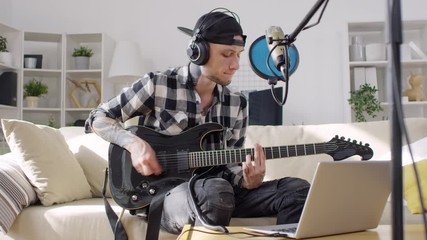 Young tattooed man playing electric guitar on sofa in the living room and singing into microphone while recording song with laptop in home studio - Powered by Adobe