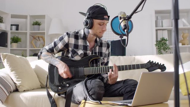 Young tattooed man playing electric guitar on sofa in the living room and singing into microphone while recording song with laptop in home studio