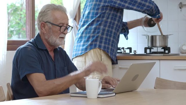 Caucasian family handsome man adult son helping his senior father solving the problems on laptop computer in the kitchen. Old man working on laptop with internet and his son do cooking in the kitchen.